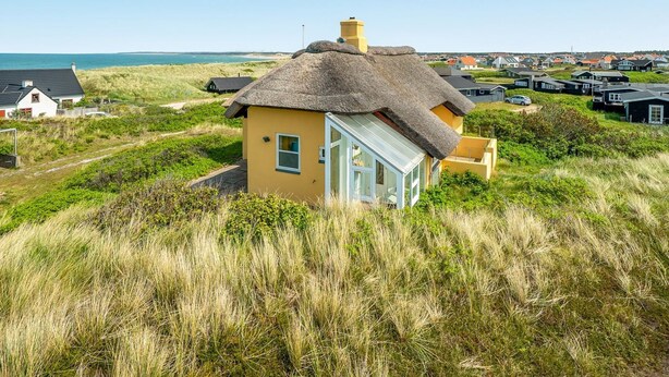 Een kusthuis met rieten dak, omgeven door gras en duinen, met uitzicht op de zee.
