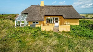 Een rieten huis met een glazen conservatorium, omgeven door weelderige begroeiing en duinen.