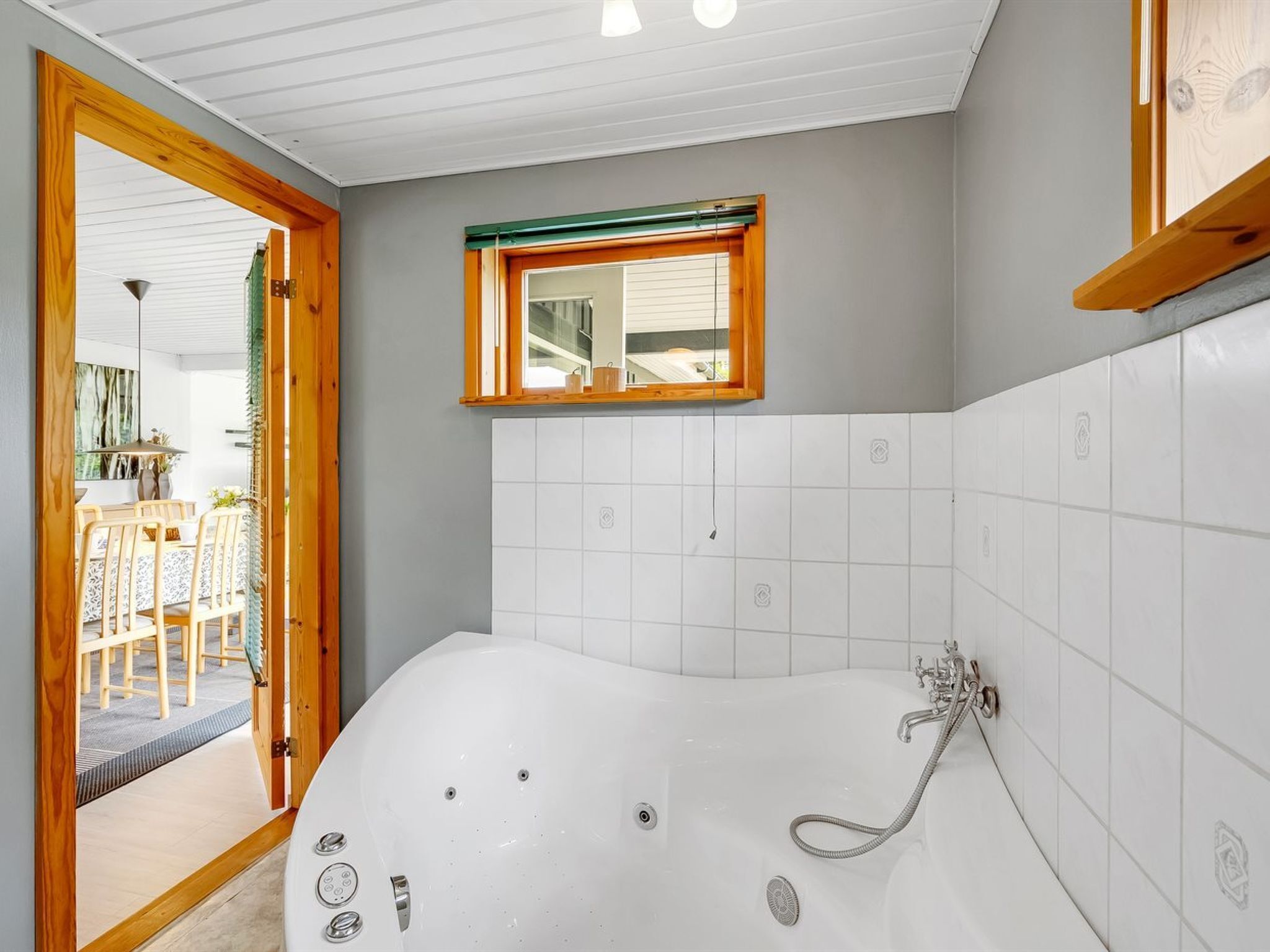 Een badkamer met een groot bad, een raam en een houten deur die leidt naar een eetruimte.