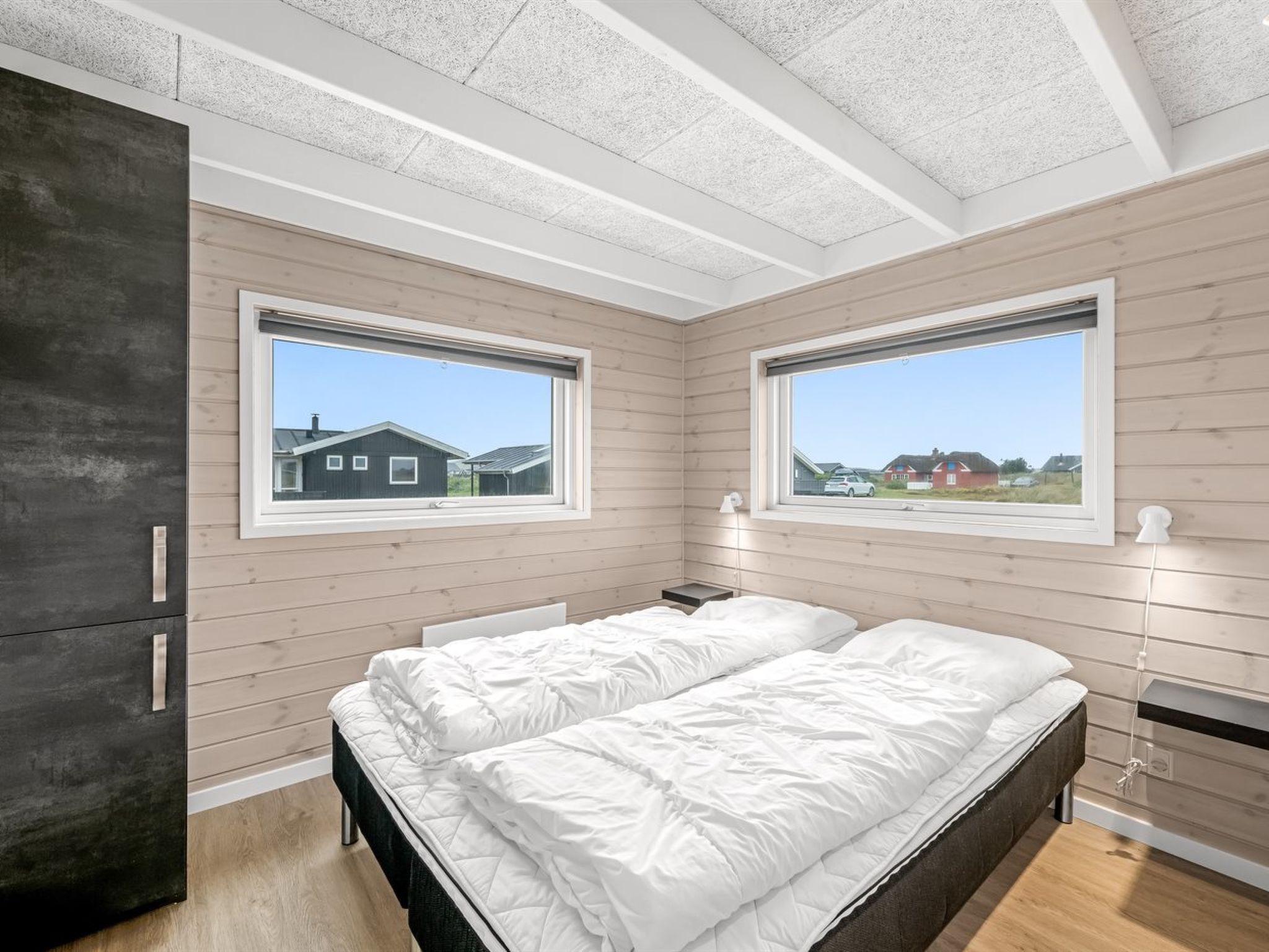 Une chambre à coucher avec un lit, une armoire et deux fenêtres donnant sur des maisons.
