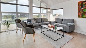 A spacious living room with a grey sectional sofa, two armchairs, and a coffee table. Large windows offer a view of the outdoors.