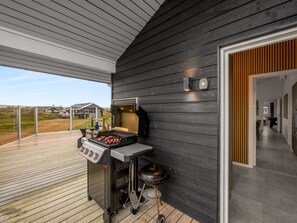 Et udendørs grillområde med indbygget ovn, gasblus og røgeovn, beliggende ved siden af et moderne hus med mørkt ydre og en træterrasse.