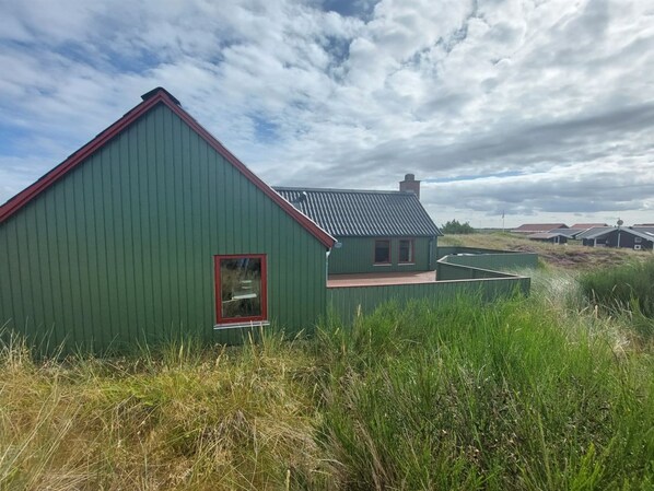 A green building with a red roof, surrounded by tall grass and other similar structures in the background.