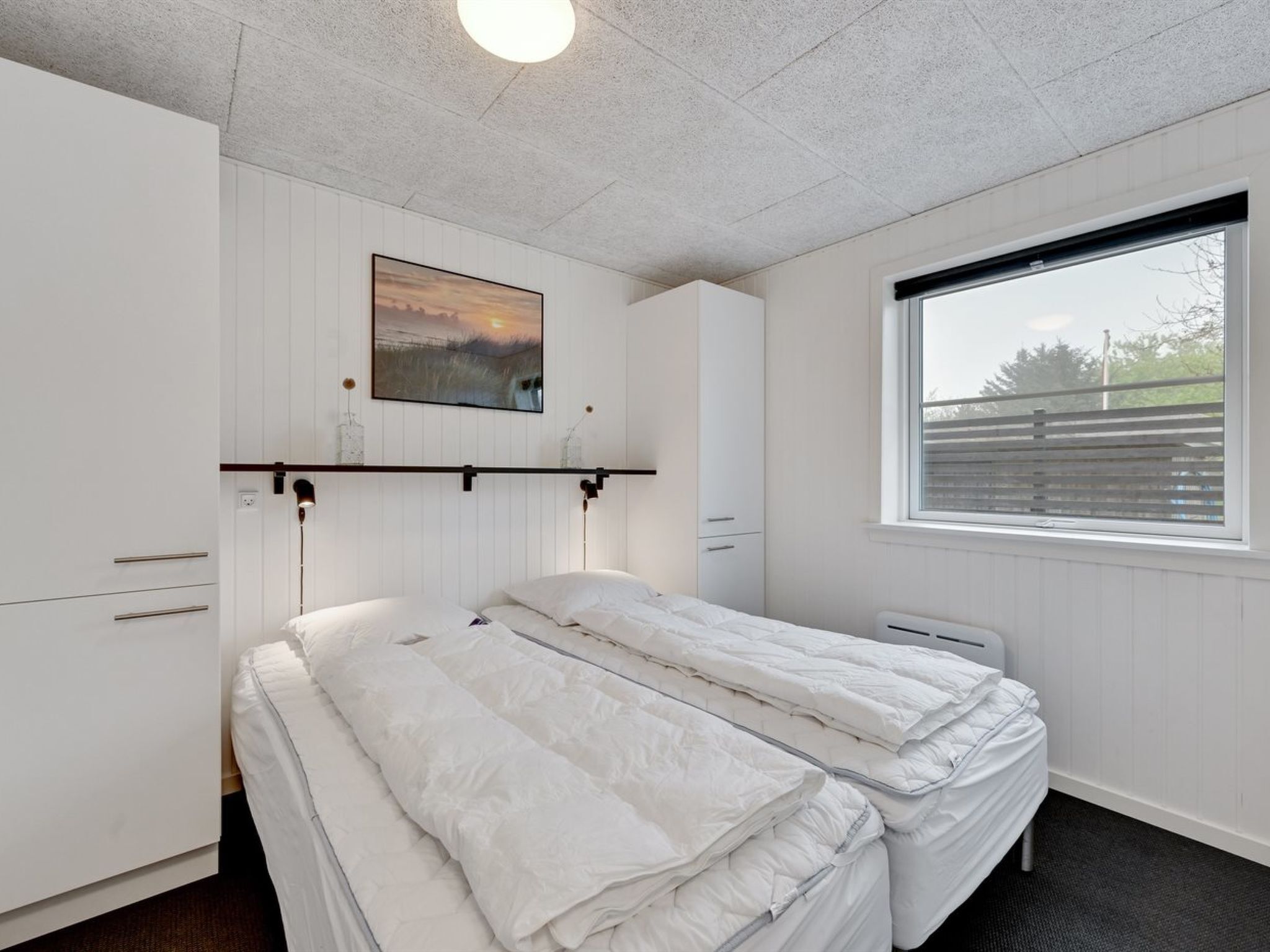Une chambre avec deux lits, une fenêtre donnant sur un beau paysage, un tableau au mur et un luminaire au plafond.