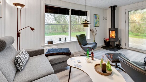 A modern living room with a sofa, a chair, a table with cards and bottles, a fireplace, and a large window with a view of the outdoors. - "Øn" -  from the sea by Interhome (Hvide Sande)