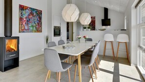 A modern dining area with a large table, grey chairs, and a wood-burning stove.