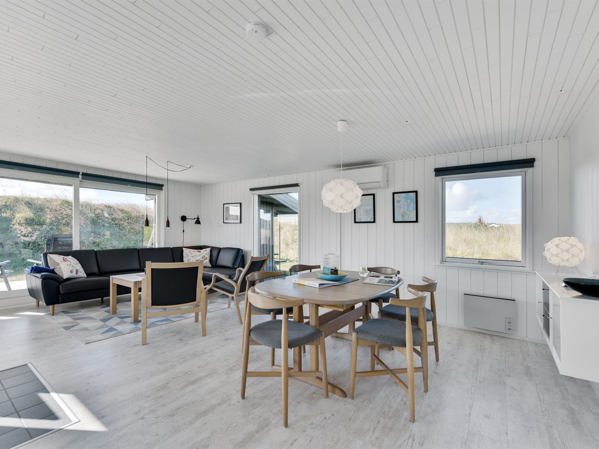 Una sala de estar moderna con un sofá negro, sillas de madera, una mesa redonda y ventanas amplias que ofrecen vistas al exterior.