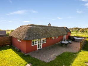 Une maison à toit de chaume avec une façade rouge, une terrasse dotée d’un jacuzzi et une clôture en bois.