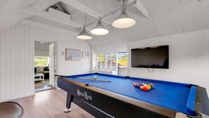 Una mesa de billar con superficie de fieltro azul, una cuesa de billar y un juego de bolas. Hay un televisor montado en la pared y una puerta que da a un área exterior.