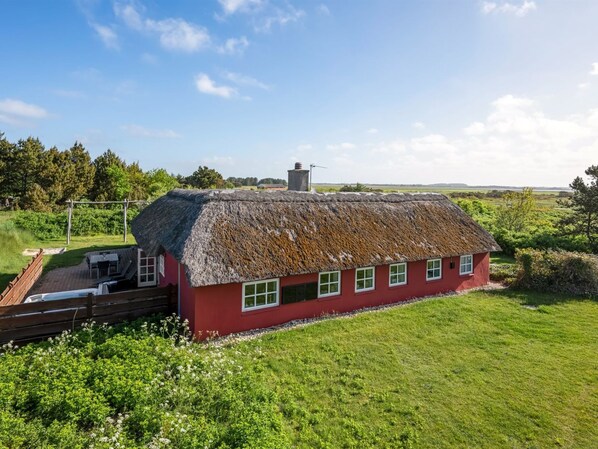 Une maison rouge avec un toit de chaume, une terrasse en bois et une cour couverte d’herbe.