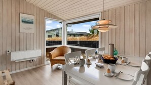 Un espace repas moderne avec une table blanche dressée pour le petit-déjeuner, une chaise en osier et une vue sur l’extérieur grâce à de grandes fenêtres.