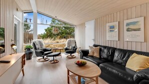 Un salon moderne avec un canapé en cuir noir, deux fauteuils, une table basse en bois sur laquelle sont disposés des fruits, et une vue sur l’extérieur grâce à de grandes fenêtres.