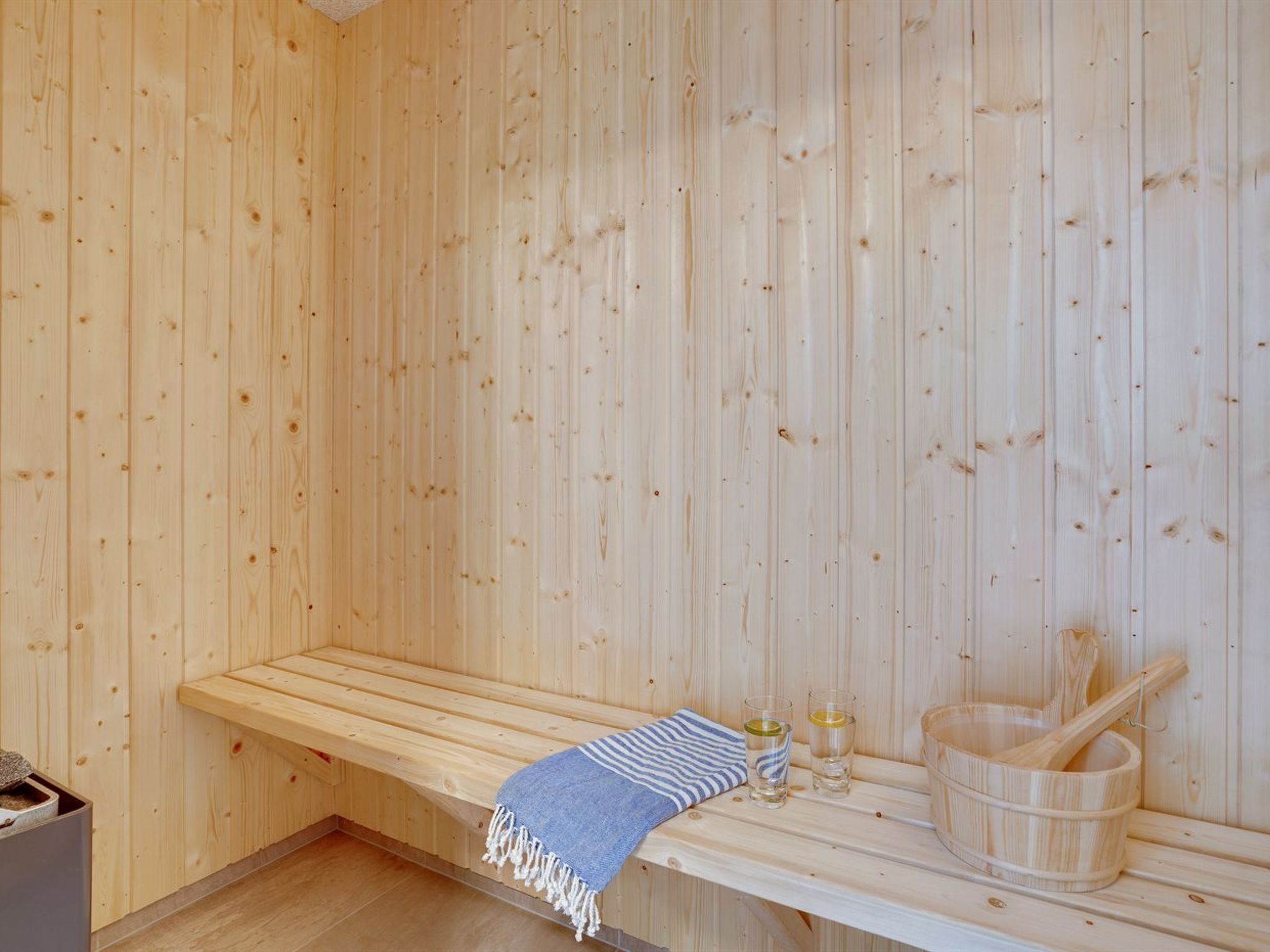 Een sauna met een houten bank, een handdoek, twee glazen en een houten emmer met een schuimspaan.