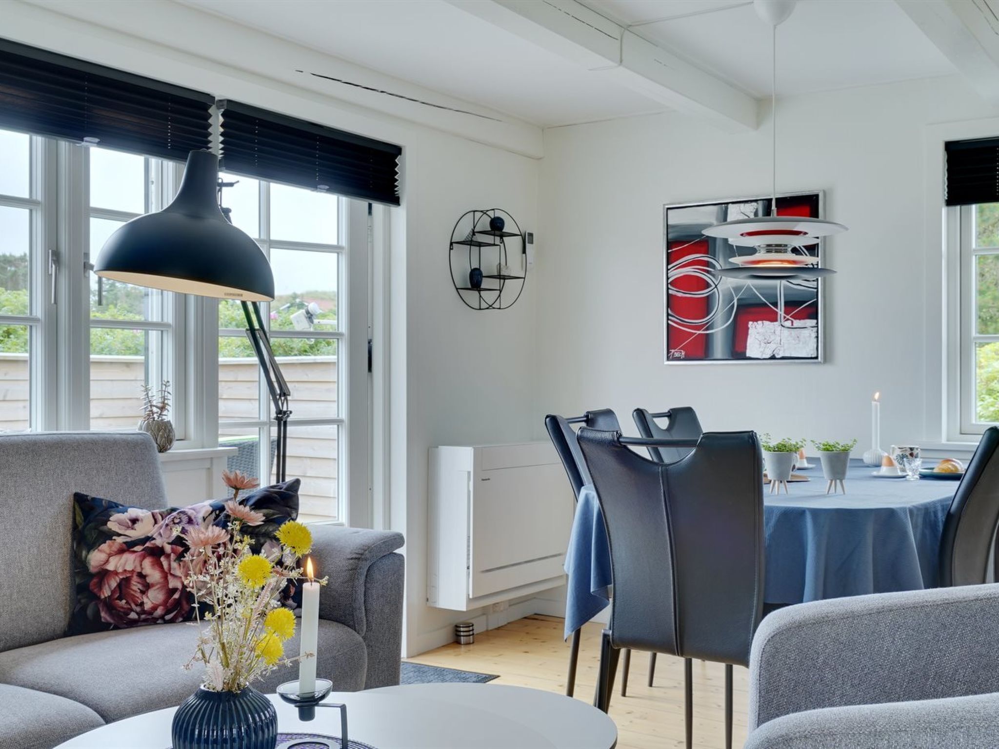 Un salon moderne comprenant un canapé, une table à manger avec des chaises, une grande fenêtre avec des stores et une œuvre d’art accrochée au mur.