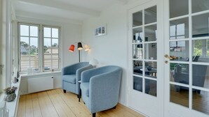 Ett rum med ett stort fönster, två grå fåtöljer, en vit dörr med glaspaneler och en golvlampa.