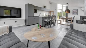 Ein modernes Wohnzimmer mit einer Couch, einem Couchtisch mit Spielkarten und einer Küchenzeile mit grauen Schränken und einem großen Fenster.