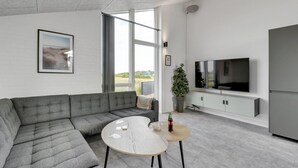 Ein modernes Wohnzimmer mit einem grauen Ecksofa, einem Holztisch, einem Flachbildfernseher und einem großen Fenster mit Blick ins Freie.