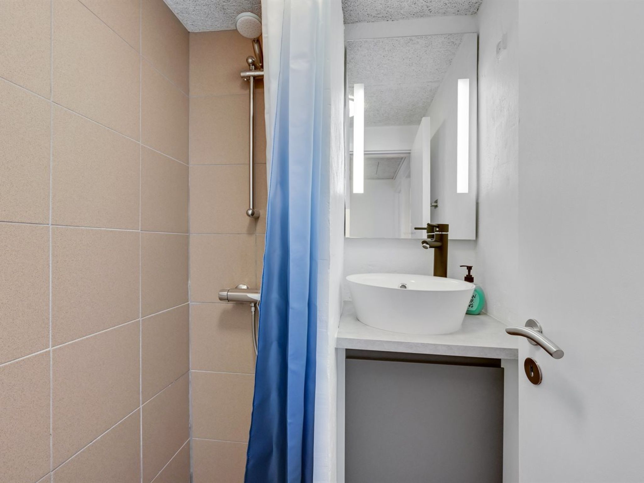 Et badeværelse med lyse beige fliser, en hvid vask med sæbedispenser, en bruser med blåt gardin og et spejl over vasken.