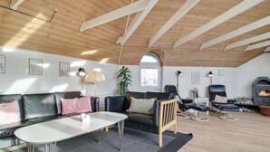 A cozy living room with a high wooden ceiling, a fireplace, a sofa, a dining table, and a large window.