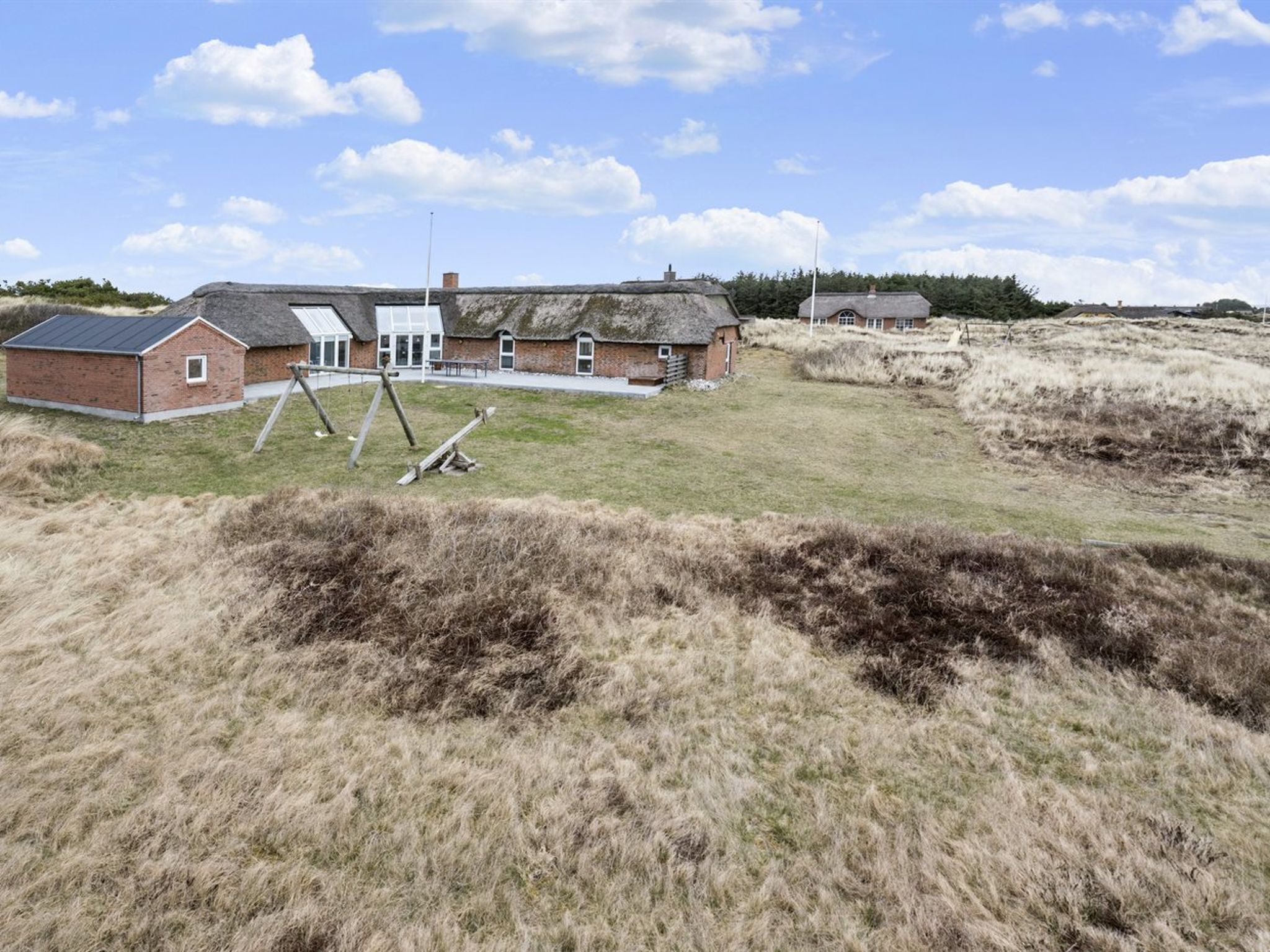 A rural setting with a large barn-like structure, a smaller building with a sloped roof, and a grassy field with a wooden swing set.