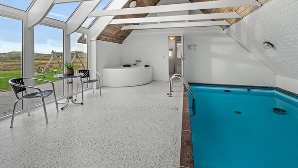 A modern indoor swimming pool with a glass ceiling, a jacuzzi, and a seating area with chairs and a table.