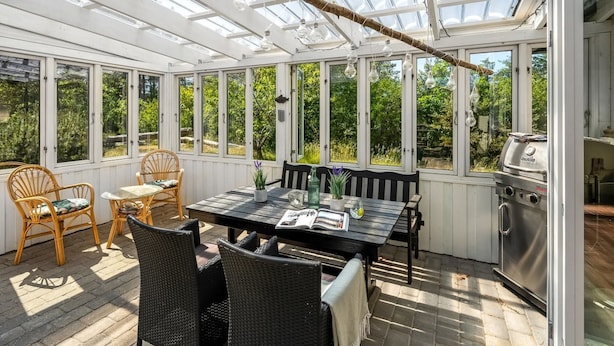 Une véranda avec une table et des chaises, un barbecue et de grandes fenêtres.