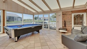 Une table de billard dans une pièce baignée de lumière, avec de grandes fenêtres, un mur en briques et un canapé en cuir.
