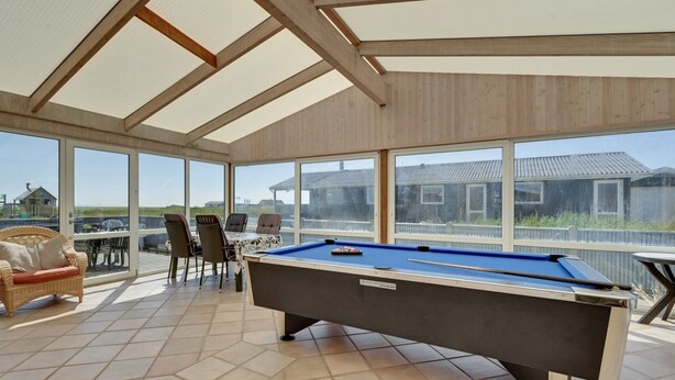 Une table de billard dans une pièce baignée de lumière, avec de grandes fenêtres, des meubles en osier et une vue sur la mer.