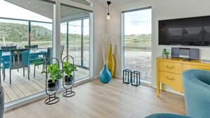 Uma sala de estar moderna com uma televisão grande, um armário amarelo e uma cadeira azul. Há duas plantas em vasos e um vaso no chão.