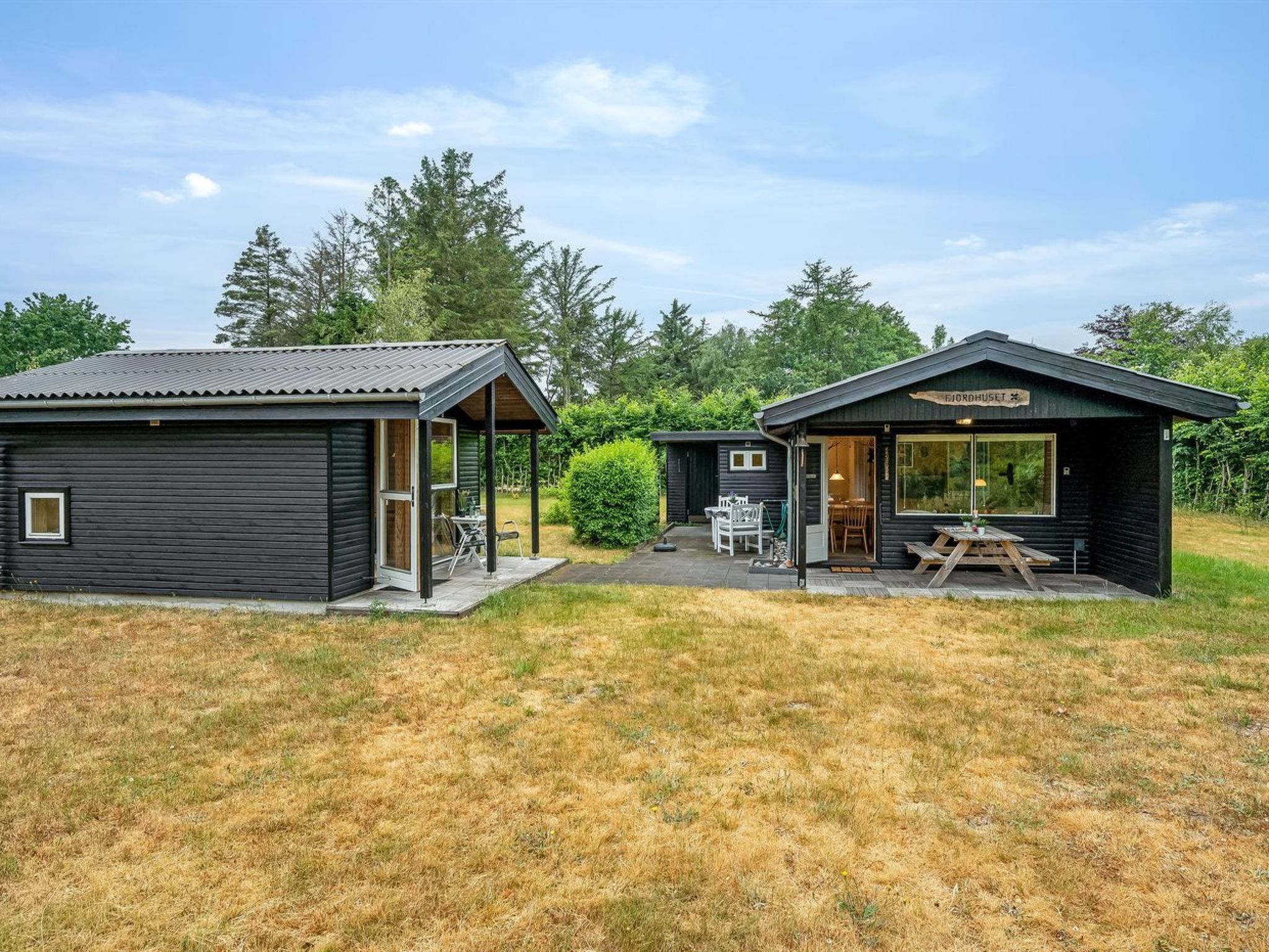 A small, dark wooden cabin with a covered patio area, a separate dining area, and a visible sign reading "Nordhuset".