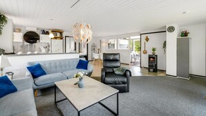 Ein modernes Wohnzimmer mit einer Couch, einem Couchtisch und einem schwarzen Ledersessel. Es gibt eine Küchenzeile mit weißen Schränken und einer hängenden Leuchte.