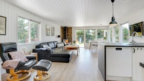A spacious living room with a black leather sofa, wooden ottoman, and a dining area with a table and chairs. A kitchen with white cabinets and a large window is visible in the background. - "Thorvor" - 1.5km from the sea by Interhome (Blåvand)