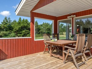 Una terraza de madera con una mesa y sillas, ventanas amplias y vistas a árboles.