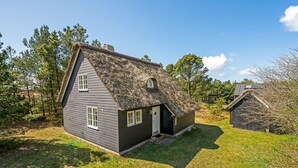 Una casa pequeña y de color oscuro con tejado de paja, rodeada de un césped y árboles.