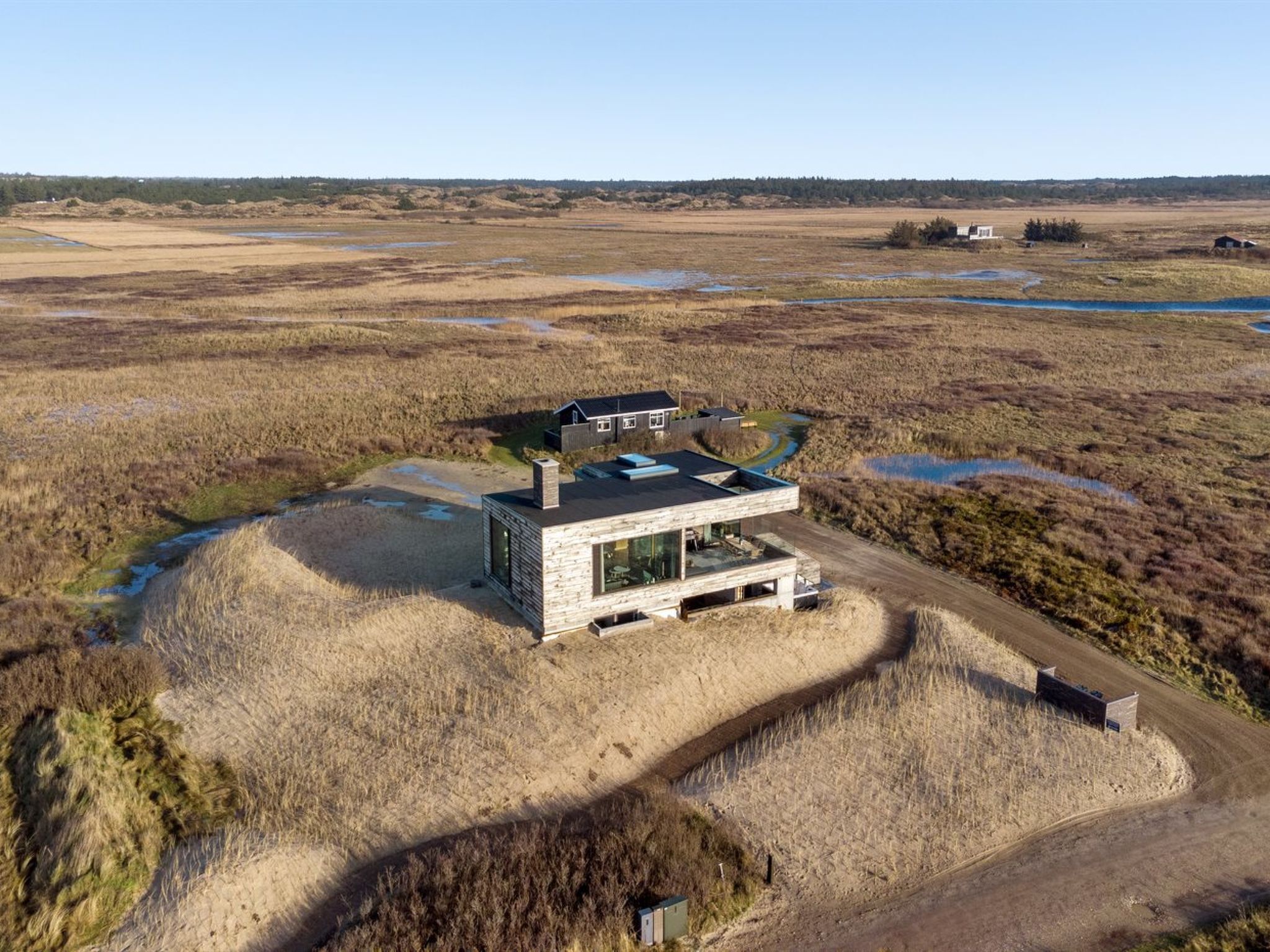 Ein modernes Haus mit flachem Dach, großen Fenstern und einer Steinaußenwand, eingebettet in eine natürliche Landschaft mit Feuchtgebieten und spärlicher Vegetation.