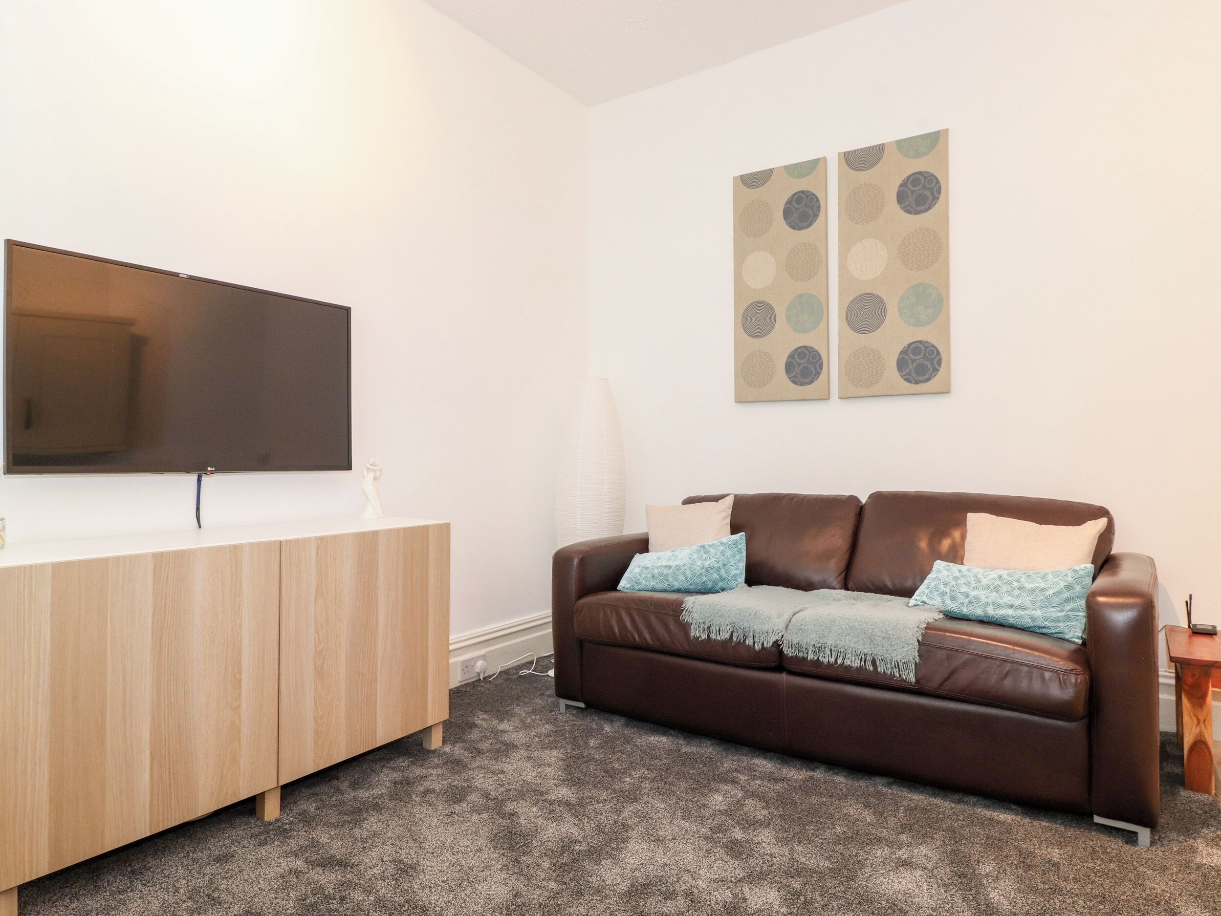 A living room with a flat-screen TV mounted on a wooden cabinet, a brown leather sofa with patterned cushions, and two abstract wall art pieces.