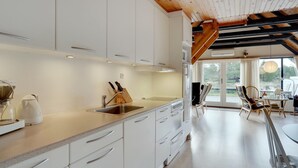 A modern kitchen with white cabinets and a wooden ceiling, leading to a dining area with a view of the outdoors.