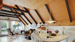 A spacious dining area with a high ceiling and exposed beams, a round table with chairs, and a view of the outdoors through large windows.
