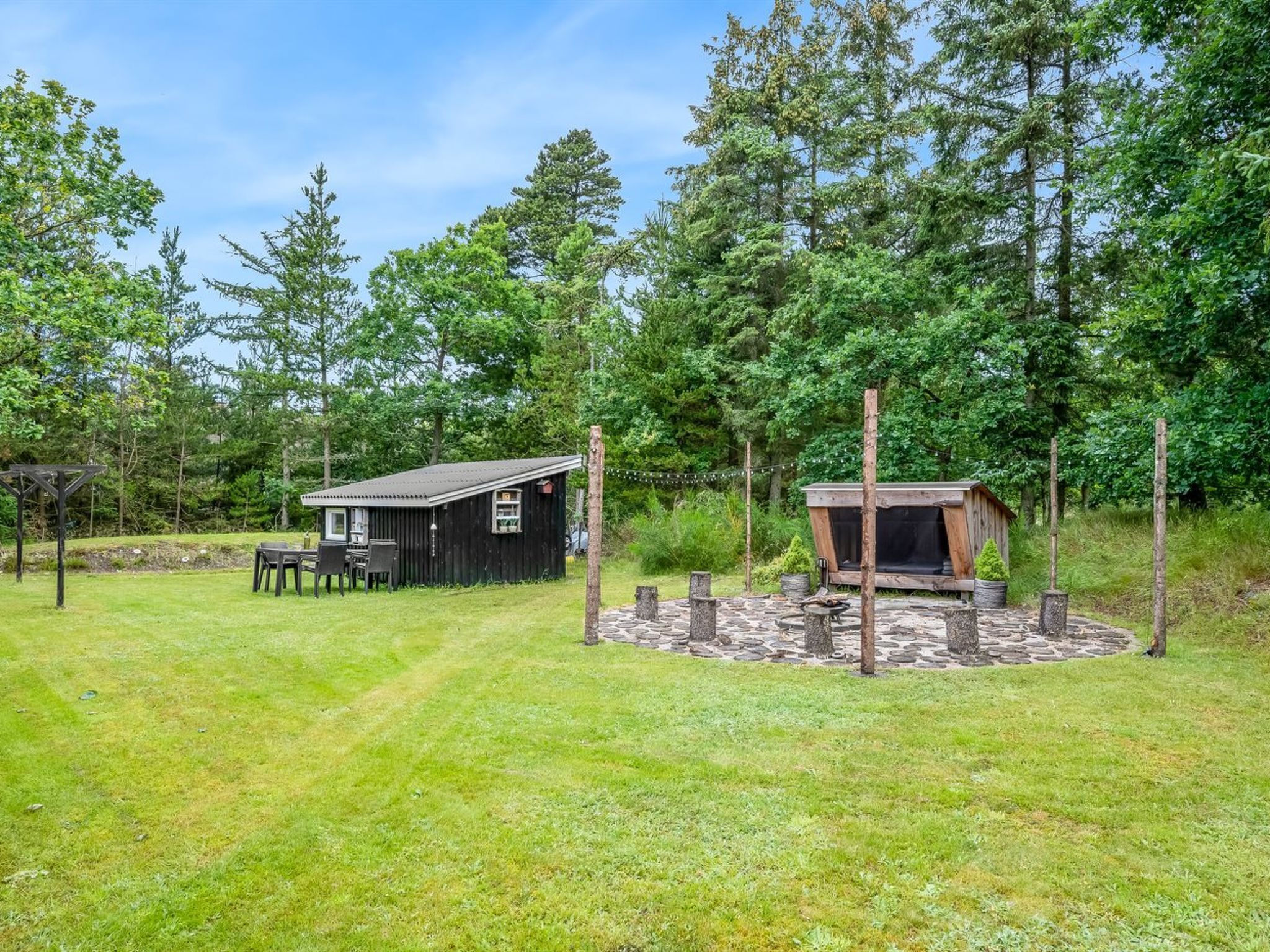A rustic cabin with a small deck and a hot tub, surrounded by a stone fire pit and a wooden pergola.