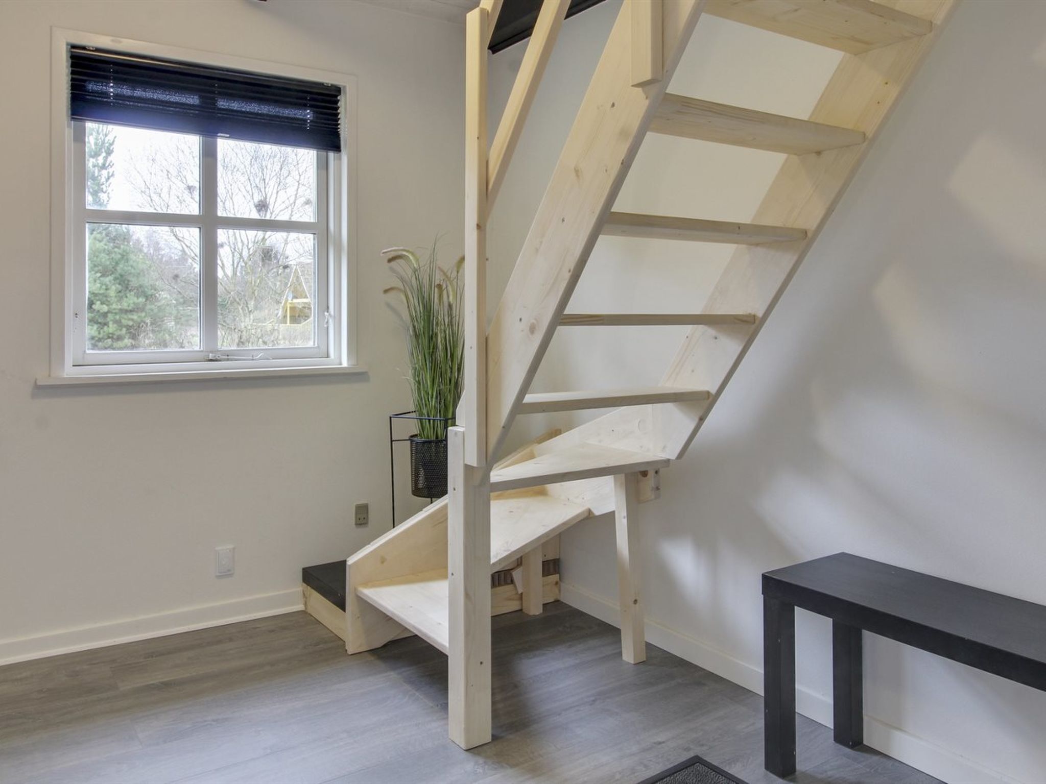 Una habitación con escalera de madera, una ventana con persianas, una planta en maceta y un banco negro.