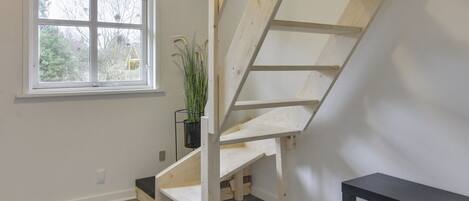 Una habitación con escalera de madera, una ventana con persianas, una planta en maceta y un banco negro.