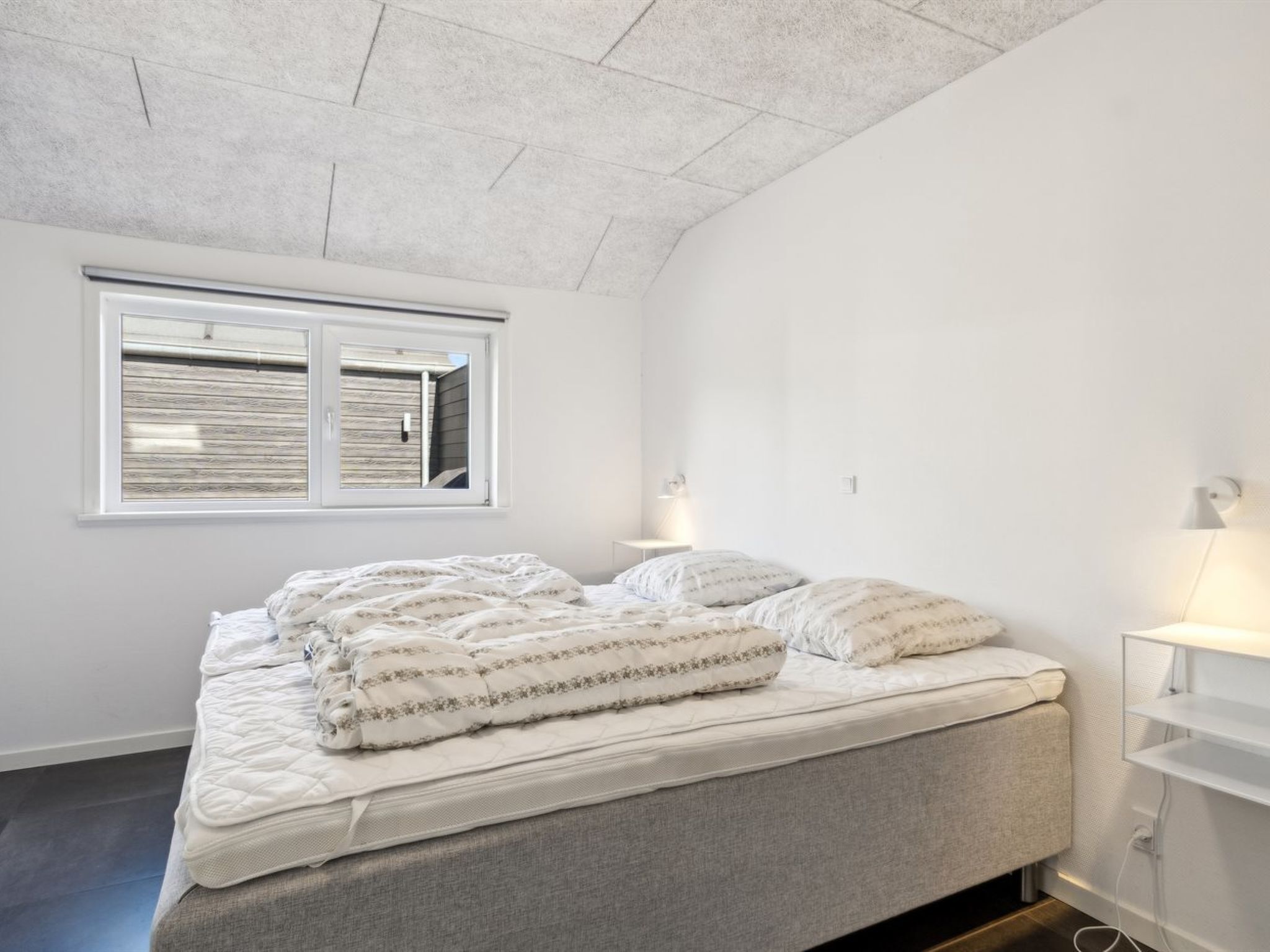 A bedroom with a bed, a window with blinds, a bedside table with a lamp, and a wall-mounted shelf.