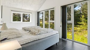 Une chambre moderne avec un grand lit, une table de chevet, une lampe et une vue sur une forêt, visible à travers de grandes portes-fenêtres coulissantes.
