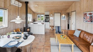 Et moderne interiør med træloft, et centralt køkkenø, et spiseområde med et rundt bord, og en opholdsstue med sofa og lænestole.