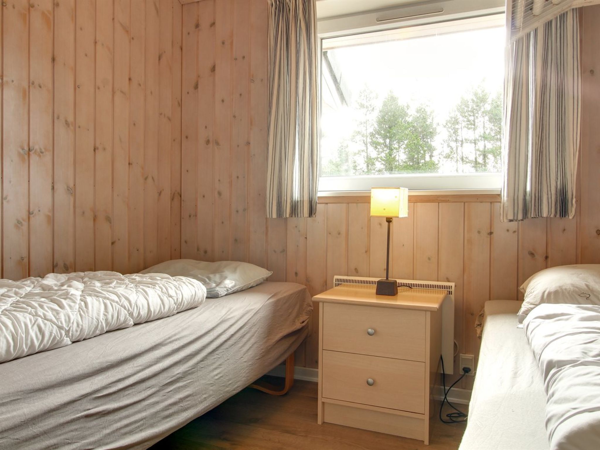 A bedroom with two single beds, a wooden nightstand with drawers, a lamp, and a window with curtains.