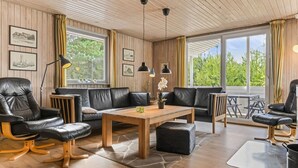 Een moderne woonkamer met een houten plafond, leren banken, een houten salontafel en grote ramen met gordijnen.