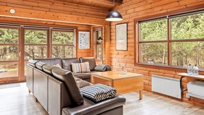 En stuga med träpanel på insidan, en soffa klädd i läder, ett matbord i trä och stora fönster med utsikt över skogen.
