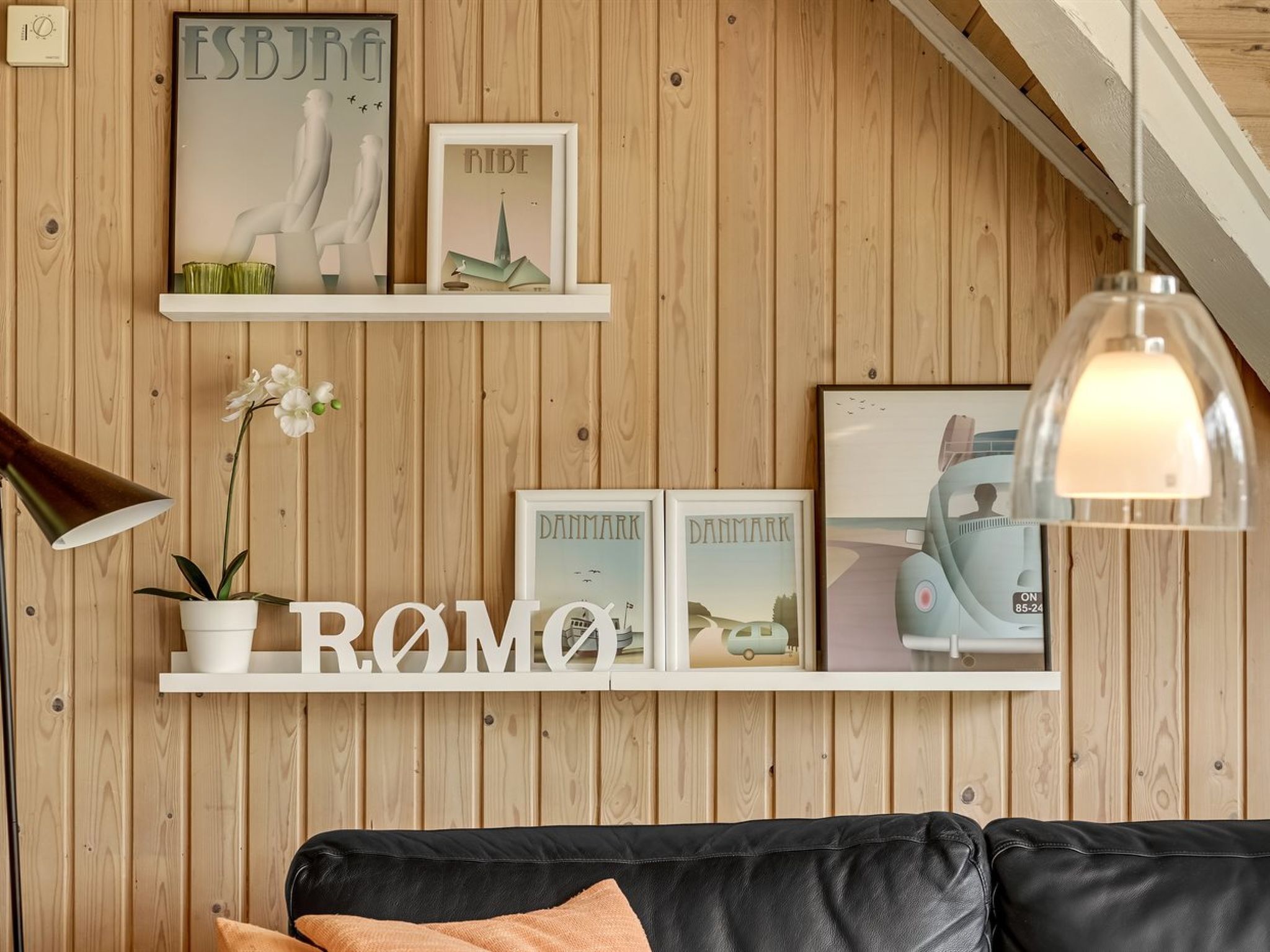 A room with wooden panel walls, a black leather sofa, a white shelf with framed pictures, a potted plant, and a hanging lamp.