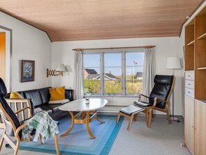 Ein Zimmer mit einer Couch, zwei Sesseln, einem Tisch, einer Lampe, einem Fenster mit Vorhängen und Blick auf Häuser.