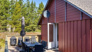A red wooden building with a satellite dish, a covered patio area with a table and chairs, and a view of trees in the background.